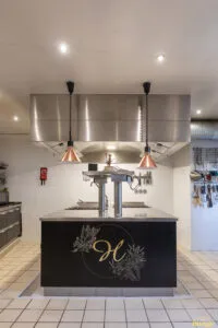 Îlot de cuisson professionnel noir et inox avec plaques et tiroirs réfrigérés, installé sous hotte en cuisine carrelée.