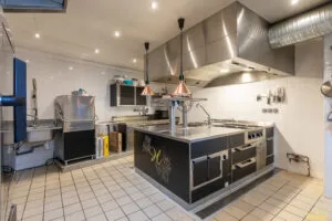 Îlot de cuisson professionnel noir et inox avec plaques et tiroirs réfrigérés, installé sous hotte en cuisine carrelée.