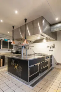 Îlot de cuisson professionnel noir et inox avec plaques et tiroirs réfrigérés, installé sous hotte en cuisine carrelée.