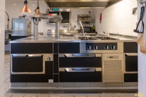 Îlot de cuisson professionnel noir et inox avec plaques et tiroirs réfrigérés, installé sous hotte en cuisine carrelée.