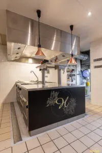 Îlot de cuisson professionnel noir et inox avec plaques et tiroirs réfrigérés, installé sous hotte en cuisine carrelée.