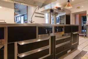 Îlot de cuisson professionnel noir et inox avec plaques et tiroirs réfrigérés, installé sous hotte en cuisine carrelée.