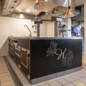 Îlot de cuisson professionnel noir et inox avec plaques et tiroirs réfrigérés, installé sous hotte en cuisine carrelée.