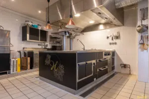 Îlot de cuisson professionnel noir et inox avec plaques et tiroirs réfrigérés, installé sous hotte en cuisine carrelée.