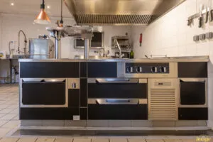 Îlot de cuisson professionnel noir et inox avec plaques et tiroirs réfrigérés, installé sous hotte en cuisine carrelée.