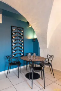 Table ronde dressée avec verres sous voûte blanche, mur bleu orné d’un porte-bouteilles vertical rempli.