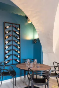 Table ronde dressée avec verres sous voûte blanche, mur bleu orné d’un porte-bouteilles vertical rempli.