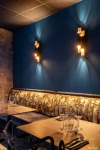 Banquette dorée capitonnée sous mur bleu avec deux appliques design allumées, table dressée et verres à vin au premier plan.