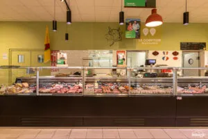 Comptoir boucherie réfrigéré vitré présentant divers morceaux de viande, vendeur derrière le comptoir, suspensions lumineuses pendantes au plafond.