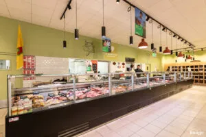Comptoir boucherie réfrigéré vitré présentant divers morceaux de viande, vendeur derrière le comptoir, suspensions lumineuses pendantes au plafond.
