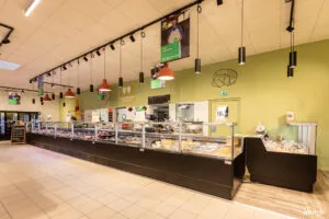 Comptoir boucherie réfrigéré vitré présentant divers morceaux de viande, vendeur derrière le comptoir, suspensions lumineuses pendantes au plafond.