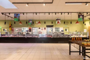 Comptoir boucherie réfrigéré vitré présentant divers morceaux de viande, vendeur derrière le comptoir, suspensions lumineuses pendantes au plafond.