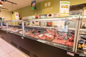 Comptoir boucherie réfrigéré vitré présentant divers morceaux de viande, vendeur derrière le comptoir, suspensions lumineuses pendantes au plafond.