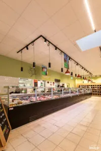 Comptoir boucherie réfrigéré vitré présentant divers morceaux de viande, vendeur derrière le comptoir, suspensions lumineuses pendantes au plafond.