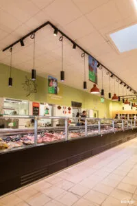 Comptoir boucherie réfrigéré vitré présentant divers morceaux de viande, vendeur derrière le comptoir, suspensions lumineuses pendantes au plafond.