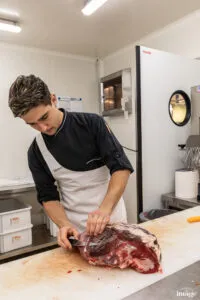 Employé en tablier découpant une grosse pièce de viande rouge sur une table de préparation.