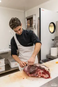 Employé en tablier découpant une grosse pièce de viande rouge sur une table de préparation.