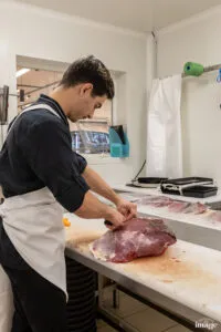 Employé en tablier découpant une grosse pièce de viande rouge sur une table de préparation.