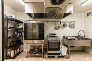 Zone de préparation : four mixte sous hotte, cuisinière et balances, étagère d’ingrédients, évier inox avec casseroles suspendues.