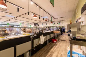 Comptoir d’épicerie : trancheuse et poste de caisse au premier plan, rayonnages garnis et employé en arrière-plan.